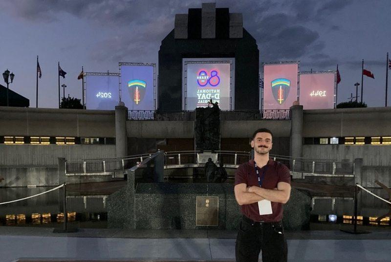 A man stands with crossed arms in front of tiers of concrete with steps, leading towards three movie screens in the distance with the words “80 YEARS NATIONAL D-DAY MEMORIAL.”